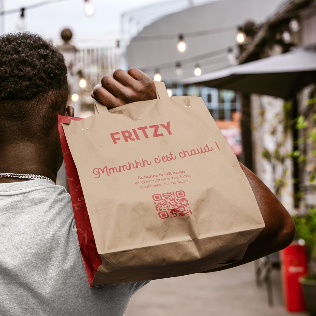 Person carrying a branded takeaway bag from Fritzy with the text 'Mmmhhh c'est chaud!' in an outdoor dining area with string lights in the background.